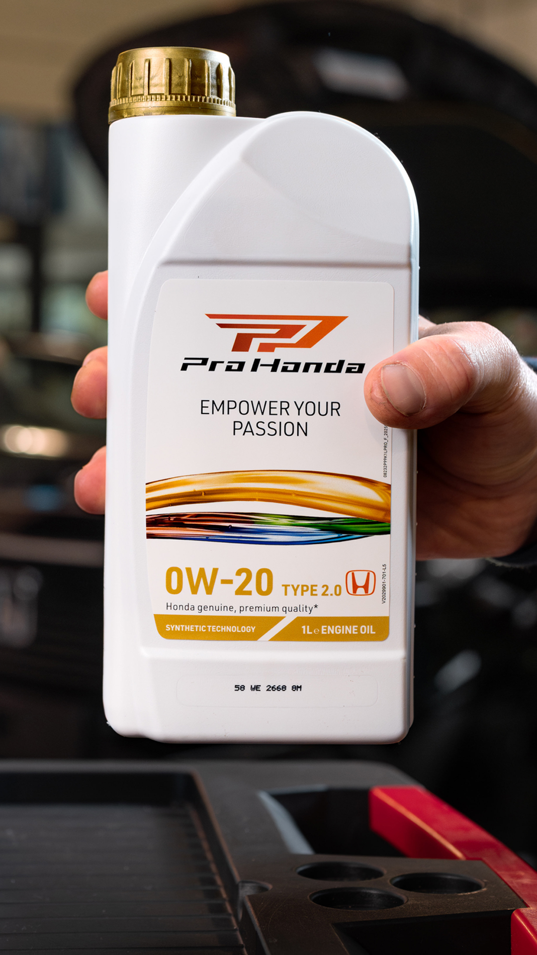 A hand holding a white plastic bottle of Pro Honda 0W-20 Type 2.0 engine oil. The bottle features the Pro Honda logo and the slogan \"EMPOWER YOUR PASSION\". The gold cap and colorful design elements are visible on the product label.