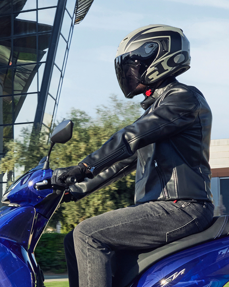 A motorcyclist wearing a black leather jacket and helmet sits on a blue Honda motorcycle. The rider is gripping the handlebars, ready to ride. In the background, a building with large windows is visible against a clear sky.