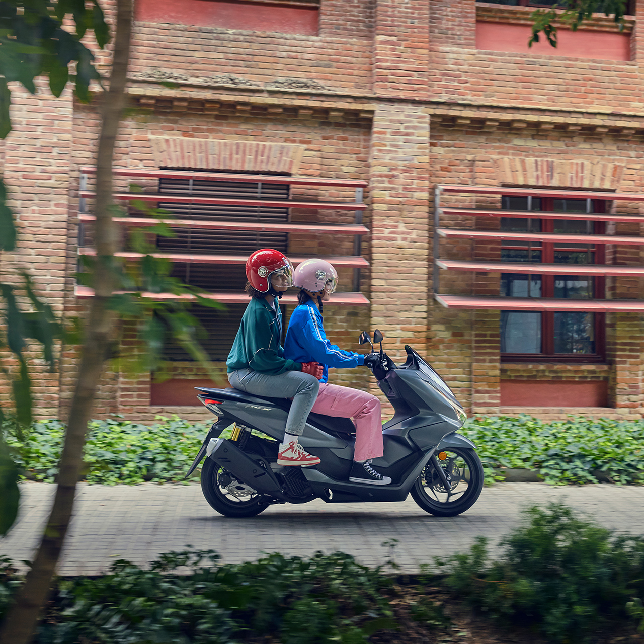 Bočný záber na figurantov pri jazde na motocykli PCX125 v meste.